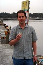 A man on a pier looking at the camera, holding in his bare hand a small silver-color fish.