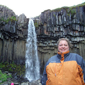 A woman, Julie Contino, standing outdoors in a windbreaker. Background is a high waterfall cascading from a high barren rockface.