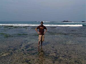 A man facing the camera, standing ankle-deep in shallow sea water.