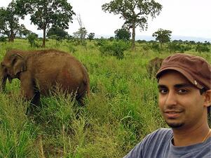 A man standing in a green grassy field looking at the camera, with a baby elephant behind him.