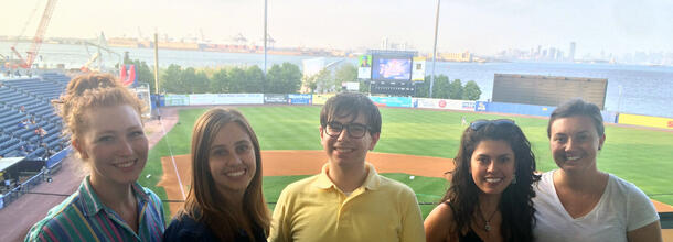 A group of five people shown in front of a baseball field.