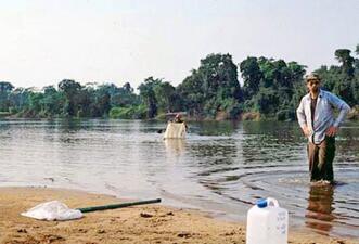 A person standing knee-deep in water. A long-handled net and a large plastic jug are on the sandy beach a few yards away.