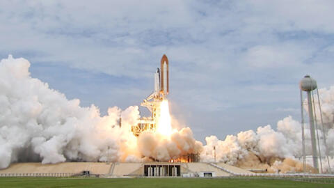 A space craft lifting off from its launch pad in daylight, with its fiery exhaust raising plumes of gas and vapor.