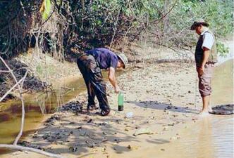 Two people wearing field clothes standing on a shallow river bank collecting specimens.