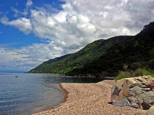 A pebbly waterfront with tree-covered hills in the background beneath a blue sky with cumulus clouds.