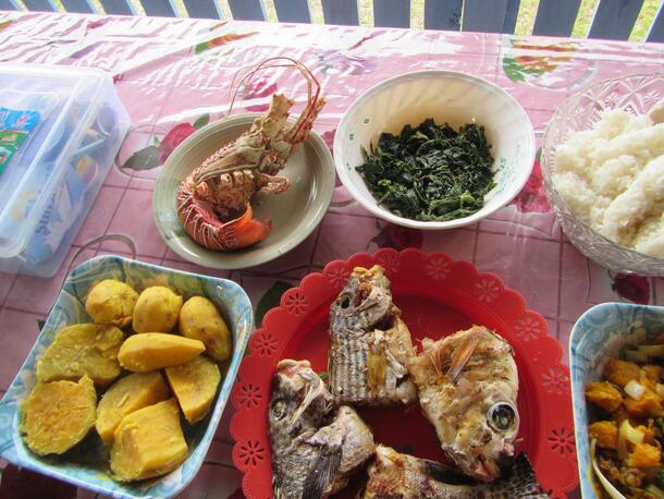A feast laid out on a table with a colorful tablecloth including plates of fish heads, greens, a lobster, starchy peeled vegetables, and rice.