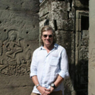 A man, Mark A. Norell, Curator and chair of the Museum's Paleontology Division, standing outdoors with stone walls in the background carved with human figures in relief.