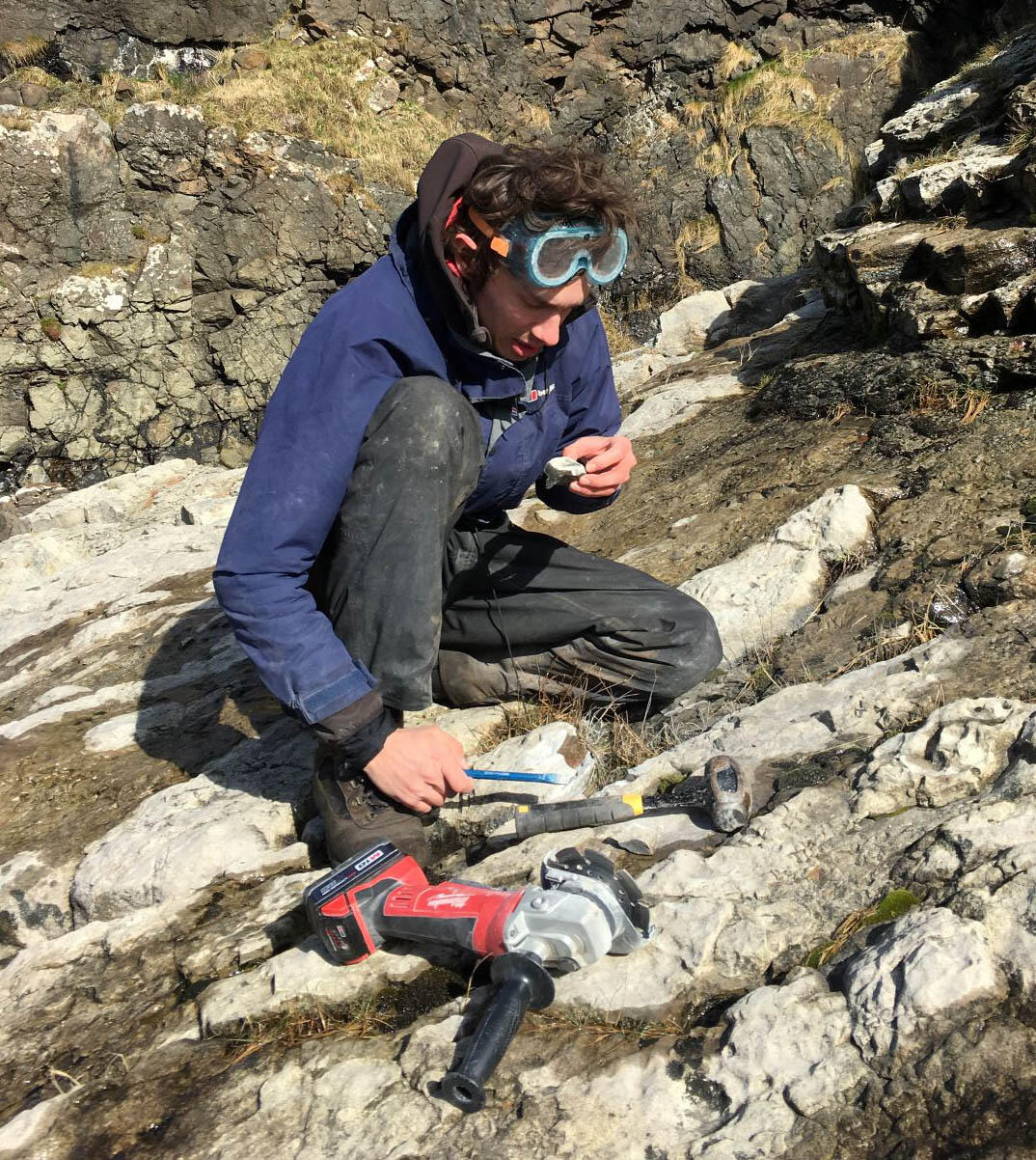 Roger Benson sits on rocky outcrop and examines a specimen in the field.