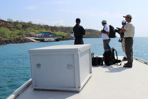 Four people stand beside a square crate on a boat deck as the boat leaves a shore.