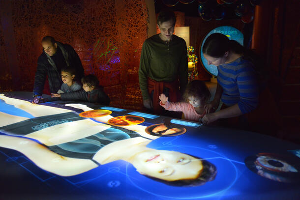 Six visitors, three adults and three children, lean over a digital interactive depicting a human body and the microbes inside.