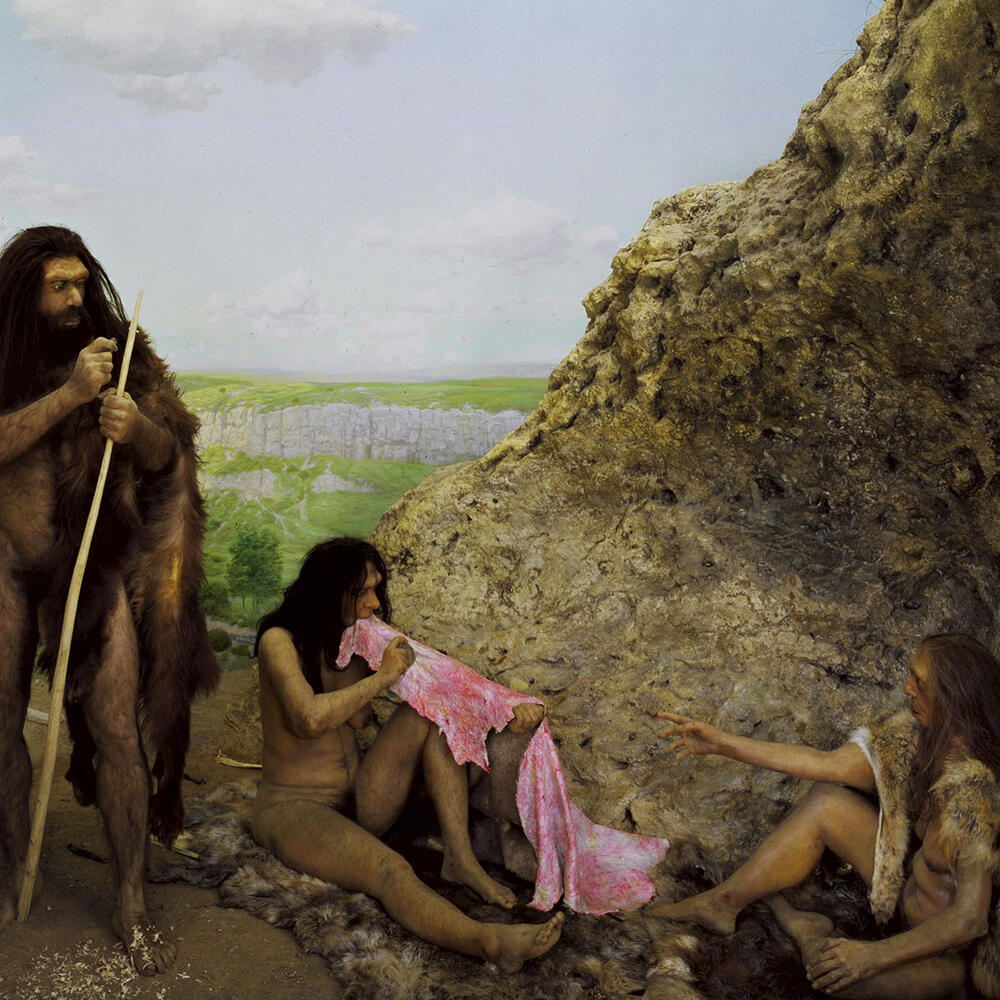 Diorama depicting three Neanderthals, one standing and holding a long stick and the others sitting and working on a piece of leather in a rocky landscape.