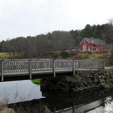 A brightly colored barn sits on the banks of a river, accessible via a wooden bridge.