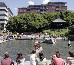 Children playing in a wading pool in a Quebec City square, surrounding by buildings with many people sitting around the pool. 