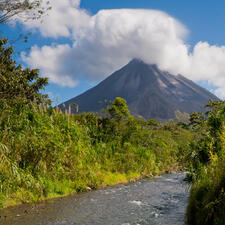 A river flanked on both sides by greenery, with a mountain the background and a bright, cloudy sky.