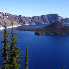 Overhead view of Crater Lake with tall trees in the foreground and mountains in the background.