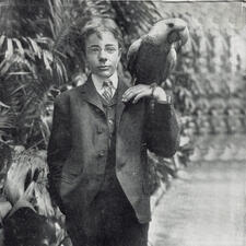Theodore Roosevelt's son Ted standing in front of plants, holding a parrot on his left hand.