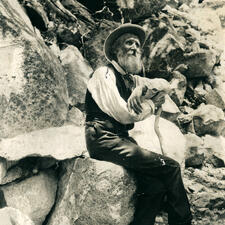 John Muir seated on a rock, holding a long stick.