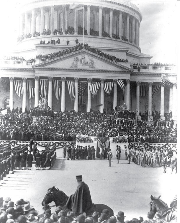 A large crowd, including someone on horseback and soldiers, assembled in front of the Capitol building which is decorated with American flags..