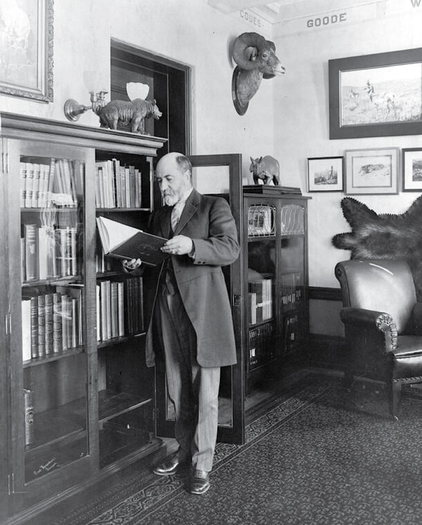 William Hornaday stands in a room in front of a bookshelf, holding a large book open.