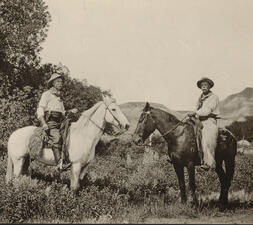 Two guides of Theodore Roosevelt's on horseback, one on a light-colored horse and the other on a dark-colored horse.