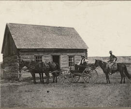 A horse-drawn carriage holding one person and another person on a house outside of an isolated ranch house.
