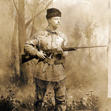 Young Theodore Roosevelt in a buckskin suit and hat, standing in a forest holding a rifle.