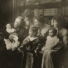 Theodore Roosevelt sits and holds a baby, with five other members of his family seated to his right.