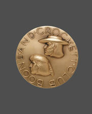 Medallion shape with text "Boone and Crockett Club" in a circle around edges and raised engravings of Daniel Boone and Davy Crockett at center.