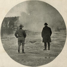 Theodore Roosevelt and John Burroughs, pictured from the back, stand in front of a Yellowstone geyser.