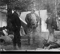 Theodore Roosevelt, John Burroughs, and another person stand around a campfire, with pitched tents behind them.