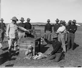 Eleven Rough Riders stand around a crate with a dark bird on top of it, and a dog sitting below.