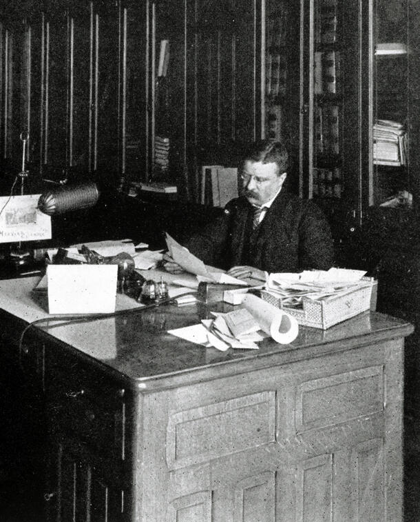 Theodore Roosevelt seated at a wooden desk, examining a piece of paper.