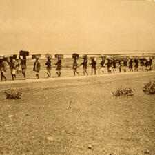 About 19 people walk in a single file line, carrying boxes and various supplies on their heads.