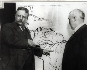 Theodore Roosevelt and another person stand in front of a map of South America, as Theodore Roosevelt points to a section of the map.