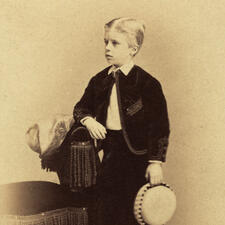 Photograph of young boy, Theodore Roosevelt, wearing dark tailored jacket and leaning one arm against a chair.