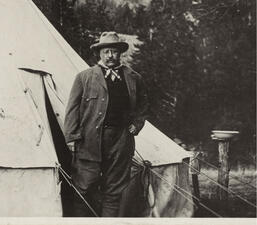 Theodore Roosevelt stands outside of a pitched tent, wearing a wide brimmed hat.