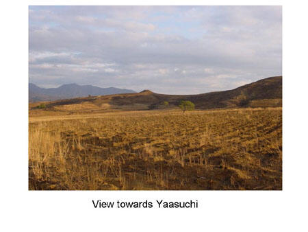 A wide flat field with dry brown grass and hills in the distance. Photo caption says "View towards Yaasuchi."