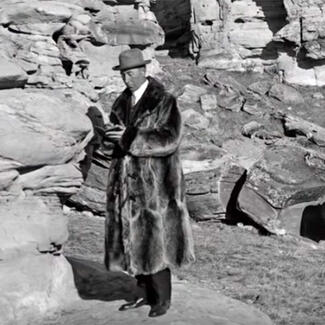Person in hat and fur coat stands in rocky, mountainous environment and holds open a small book.
