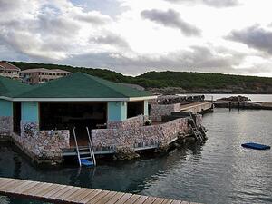 A low building on a pier in a wide lake with green hills along the shore.