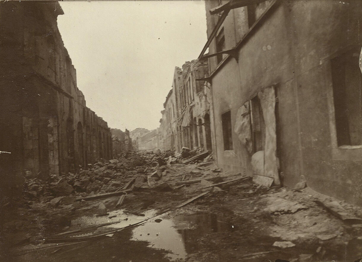 A narrow street with debris piled up in the middle and flanked by buildings in ruin.