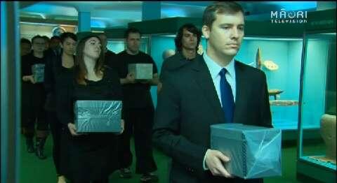 A procesion of people walk past museum displays, each carrying a box in their hands.