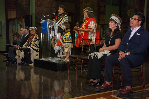 An adult and child in intricate, fringed blankets stand at a podium as seven other people sit or stand beside them on a stage.