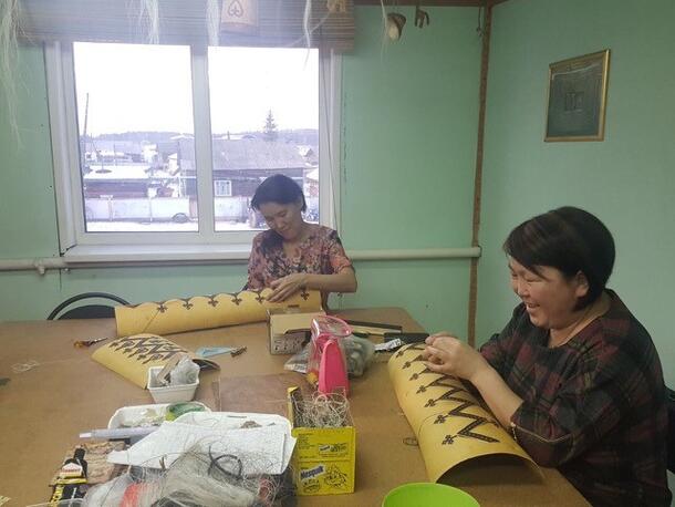 Two women sit at a table and paint designs on bark.