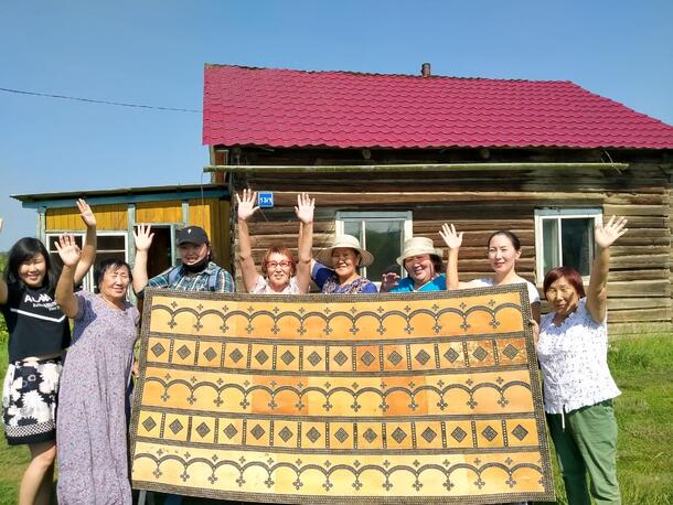 Eight people stand outside together, holding a large painted birchbark panel and waving.