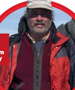 Outdoor close-up of man with short beard looking at camera. Wearing layers of warm clothes: red jacket; baseball cap shades eyes from sun.