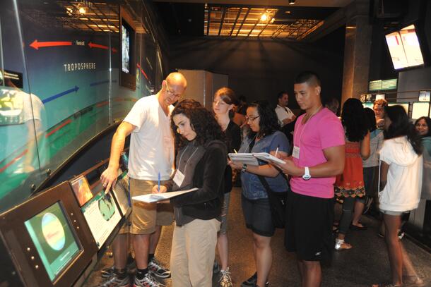 Several young adults writing in notebooks in a Museum exhibition hall as another adult points to  a map in a section about the troposphere.